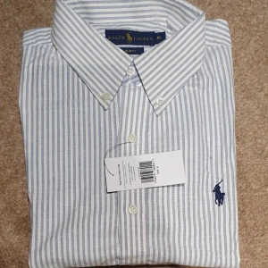 Randig skjorta från Ralph Lauren - Klassisk skjorta från Ralph Lauren i vitt och ljusblått med smala ränder. Skjortan har button down-krage, broderad logga på bröstet och knäppning framtill. Tillverkad i mjuk bomull och passar perfekt till en clean och stilren look.