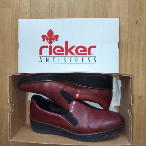 Vinröda skinnloafers från Rieker - Snygga vinröda loafers från Rieker i skinn med elastiska partier på sidorna för enkel påtagning. Skorna har en platt, svart sula med bra grepp och en klassisk rund tå. Perfekta för dig som vill ha både stil och komfort. Nypris:700, använd fåtal gånger.