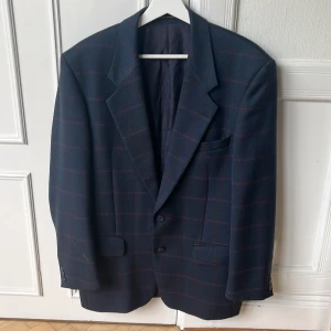 Blå rutig kavaj med röda detaljer - Snygg blå kavaj med rött rutmönster och klassisk passform från Yves Saint Laurent. Kavajen har tre knappar vid ärmslutet, fickor med lock och en bröstficka. Fodret är ljust och mjukt, perfekt för dig som gillar stilrena och tidlösa plagg. Armlängd: 61, Axelbredd: 54