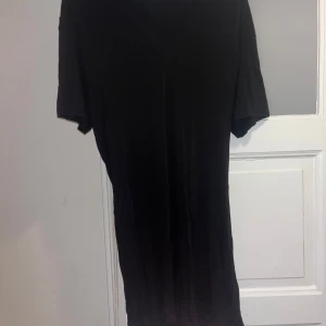 Svart långklänning med v-ringning - Svart långklänning med v-ringad hals och korta ärmar. Klänningen har en vit knytdetalj i midjan som ger en snygg siluett. Enkel och stilren design, perfekt för dig som gillar minimalistiskt mode.