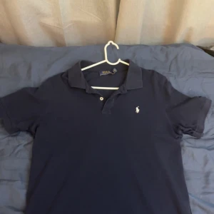 Mörkblå pikétröja från Polo Ralph Lauren - Klassisk mörkblå pikétröja från Polo Ralph Lauren med vit broderad logga på bröstet. Tröjan har korta ärmar, krage och knäppning med två knappar. Perfekt för en clean och stilren look.