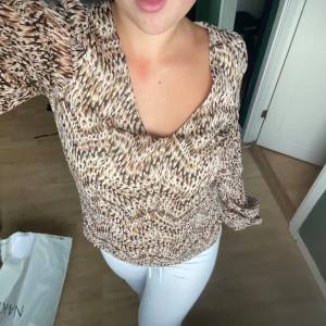 Brunmönstrad blus med v-ringning - Säljer en snygg blus med v-ringning och långa ärmar. Blusen har ett brunt och beige mönster som påminner om djurprint och är tillverkad i ett lätt, flowy material. Ärmarna har en liten knapp vid handleden för extra detalj. perfekta höstfärger som passar fint i höstgarderoben!!