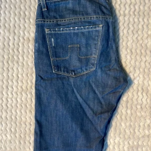 Blå jeansbyxor från J.Lindeberg - Snygga blå jeansbyxor med slitningar från J.Lindeberg med klassisk femficksdesign och raka ben. Byxorna har en diskret logga i metall på bakfickan och ljusa slitningar framtill för en trendig look. Perfekta för dig som gillar stilrena och tidlösa jeans.