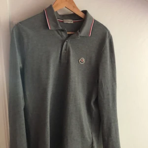 Grå långärmad pikétröja från Moncler - Säljer en stilren grå långärmad pikétröja från Moncler. Tröjan har klassisk krage med röd, vit och blå rand samt matchande detaljer vid ärmslut. Bröstet pryds av Moncler loggan Perfekt för en clean look!🔥
