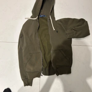 Olivgrön hoodie från Polo Ralph Lauren - Säljer en olivgrön hoodie från Polo Ralph Lauren med dragkedja och huva. Jackan har två fickor framtill, ribbade muddar och en liten broderad logga på bröstet. Perfekt för en avslappnad stil.