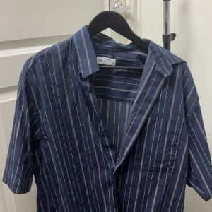 Blå randig skjorta från Zara - Snygg blå skjorta från Zara med vita ränder och korta ärmar. Skjortan har klassisk krage och bröstficka, perfekt för en avslappnad stil. Tillverkad i mjukt material som känns skönt mot huden.