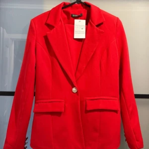 Röd kostym med kavaj och byxor från Vesper Chicy - Snygg röd kostym med klassisk kavaj och matchande byxor. Kavajen har slag, två fickor med lock och silverfärgade knappar. Byxorna är raka med knapp och hällor i midjan. Perfekt för dig som vill sticka ut med färg.