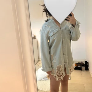 Ljusblå oversized jeansjacka med slitningar - Säljer en ljusblå oversized jeansjacka med stora slitningar och fransar längs nederkanten. Jackan har långa ärmar, bröstfickor med lock och knäppning framtill. Perfekt för en avslappnad och trendig look.