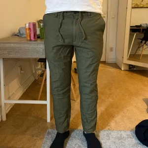 Olivgröna linnebyxor med snörning - Säljer ett par olivgröna linnebyxor med snörning i midjan. Byxorna har normal passform och är tillverkade i bomull med lite elastan för extra komfort. Perfekta för en avslappnad och stilren look.