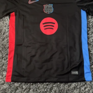 FC Barcelona svart fotbollströja Lamine Yamal 19 - Säljer en svart FC Barcelona fotbollströja från Nike med Lamine Yamal och nummer 19 på ryggen. Tröjan har röda och blå detaljer på sidorna, rött Spotify-tryck på bröstet och klubbmärke framtill. Perfekt för dig som älskar Barca!