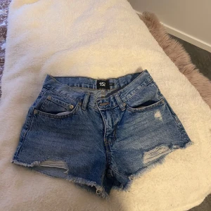 Blå lågmidjade  jeansshorts  - Säljer ett par blå lågmidjade jeansshorts från lager 157 med fransiga benslut och slitna detaljer framtill. Klassisk femficksmodell med bälteshällor och knappgylf. Perfekta för varma dagar.
