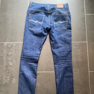 Blå jeans från Nudie Jeans - Säljer ett par klassiska blå jeans från Nudie Jeans med raka ben och snygga kontrastsömmar. Byxorna har fem fickor och läderpatch bak i midjan. Perfekta för dig som gillar stilrena och tidlösa jeans.