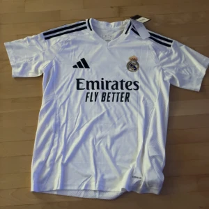 Real Madrid fotbollströja Bellingham 5 - Säljer en vit Real Madrid fotbollströja från Adidas med svarta detaljer och Bellingham 5 på ryggen. Tröjan har korta ärmar, klubbmärke på bröstet och sponsortryck framtill. Perfekt för fans av laget.