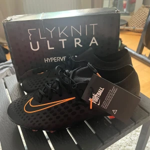 Nike Hypervenom Flyknit Ultra fotbollsskor - Svarta Nike Hypervenom Flyknit Ultra fotbollsskor med orange swoosh och mönstrad ovandel. Skorna har snörning och en platt sula med dobbar i orange och guld. Perfekta för dig som vill sticka ut på planen.