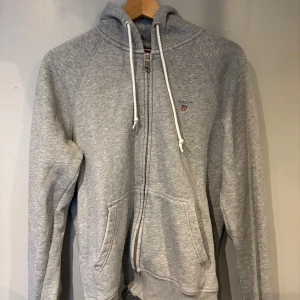 Grå hoodie med dragkedja från GANT - Säljer en klassisk grå hoodie från GANT i storlek M. Tröjan har dragkedja, huva med snören och två fickor framtill. Broderad GANT-logga på bröstet. Perfekt för en avslappnad stil.