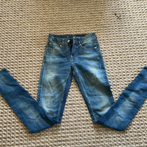 Blå jeans från Tiger of Sweden - Säljer ett par klassiska blå jeans från Tiger of Sweden i modellen Slight. Jeansen har normal passform, raka ben och snygga slitningar framtill. Tillverkade i Italien och perfekta för dig som gillar stilrena jeans med en modern touch.