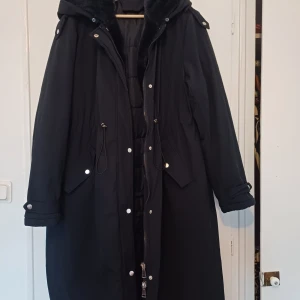 Svart lång parkajacka med huva - Säljer en svart lång parkajacka med huva och dragsko i midjan. Jackan har dragkedja och knappar framtill samt fickor med lock. Perfekt för kyliga dagar och har en klassisk stil.