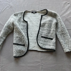 Grå cardigan jacka med svarta detaljer från Bik Bok - Säljer en gråvit, grovstickad cardigan jacka från Bik Bok med svarta kanter och dekorativa fickor. Koftan är öppen framtill och har en kort, croppad passform. 