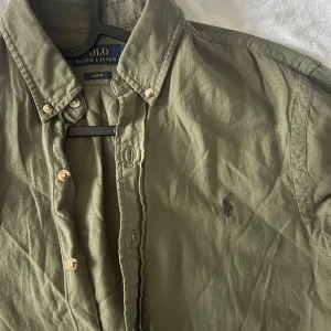 Olivgrön skjorta från Polo Ralph Lauren - Snygg olivgrön skjorta från Polo Ralph Lauren med klassisk button-down krage och broderad logga på bröstet. Skjortan har långa ärmar och knäppning framtill. Perfekt för en stilren look.