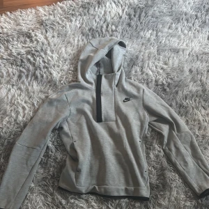 Grå Nike tech fleece hoodie - Säljer en stilren grå hoodie från Nike med halv dragkedja framtill och huva. Tröjan har lång ärm och är perfekt för en sportig look. Nike-logga på bröstet och mjukt material.