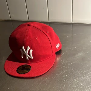 Röd New Era keps med NY-logga - Säljer en röd keps från New Era med vit broderad NY-logga framtill. Klassisk 59FIFTY-modell med platt skärm och New Era-logga på sidan. Perfekt för dig som gillar sportig stil.