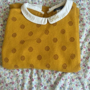 Gul stickad tröja med krage från Okaidi - Superfin gul stickad tröja från Okaidi med glittriga prickar och vit krage där det står 'Smile' broderat. Tröjan har långa ärmar och en söt, klassisk look med knapp i nacken. Perfekt för dig som gillar färg och detaljer!
