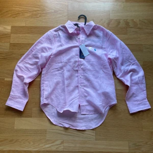 Ljusrosa linneskjorta från Polo Ralph Lauren - Säljer en ljusrosa skjorta från Polo Ralph Lauren i 100% linne. Skjortan har klassisk krage, knäppning framtill och broderad logga i blått på bröstet. Perfekt för en stilren och avslappnad look.