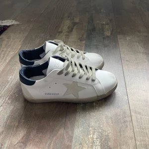 Vita sneakers från Golden Goose - Snygga vita sneakers med grå stjärna på sidan och grå snörning. Skorna har mörkblå häl och klassisk rund tå. Perfekta för en stilren och avslappnad Grisch look