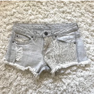 Grå jeansshorts med spetsdetalj från Bershka - Superfina grå jeansshorts från Bershka med fransig kant och snygga slitningar. Ena framsidan har en vit spetsdetalj som ger shortsen en unik look. Perfekta för varma dagar och en trendig stil.