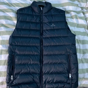 Svart dunväst från Gant - Snygg svart dunväst från Gant med dragkedja framtill och två fickor med dragkedja. Västen har en klassisk quiltad design och hög krage. Perfekt för lager-på-lager under kyliga dagar. Köptes ny för 2000 kr