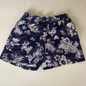 Blå blommiga badshorts från Polo Ralph Lauren - Mörkblå badshorts med vitblommigt mönster och ananastryck från Polo Ralph Lauren. Shortsen har snörning i midjan och innerbyxa i mesh. Perfekta för stranden eller poolen.