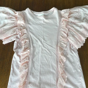 Ljusrosa volangtopp från Sofie Schnoor - Supersöt ljusrosa topp med korta volangärmar och broderade spetsdetaljer från Sofie Schnoor. Toppen har dekorativa volanger framtill och är tillverkad i mjuk bomull. Perfekt för en feminin och lekfull stil.