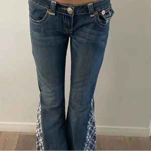 Blå jeans med spetsdetaljer - Snygga blå jeans från True Religion med unika spetsdetaljer längs benen. Byxorna har en bootcut-stil och är lågmidjade. De har också dekorativa sömmar och knappar på bakfickorna. Säljer dessa byxor då de var för små för mig, de är köpt här på Plick💗(Inte mina bilder) bara att skriva om man har några funderingar!💕