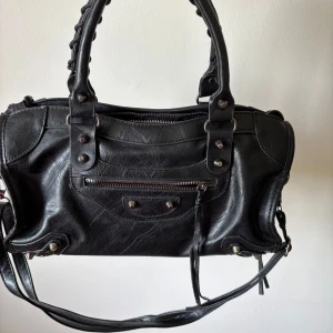 Svart axelväska i skinn med detaljer - Säljer en svart axelväska i skinn med både handtag och axelrem. Väskan har dragkedja upptill, dekorativa nitar och dragkedjeficka framtill samt långa skinnband som detaljer. Kan mötas upp i centrala Stockholm!