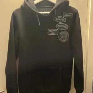 Svart hoodie från Mercier med patch-detaljer - Svart hoodie från Mercier med flera patchar och broderade loggor på bröstet. Tröjan har huva med dragsko och en stor magficka framtill. Perfekt för en avslappnad och sportig stil.