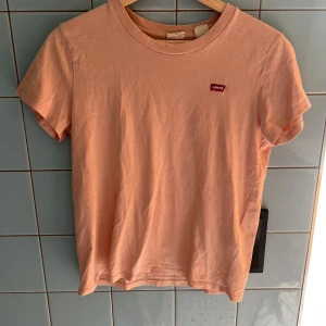 Ljusrosa t-shirt från Levi's - Säljer en ljusrosa t-shirt från Levi's med klassisk rund hals och liten röd Levi's-logga på bröstet. T-shirten är i mjuk bomull och har korta ärmar. Perfekt för en avslappnad stil.