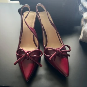 Vinröda pumps med rosett och spetsig tå - Säljer ett par glansiga vinröda pumps med öppen häl och smal klack. Skorna har en snygg rosett framtill och spetsig tå, vilket ger en elegant look. Perfekta för dig som vill sticka ut med färg och stil. Oanvända.
