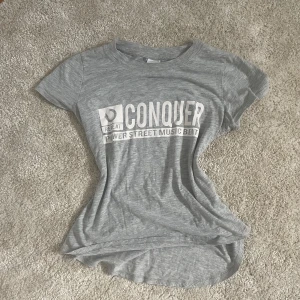 Grå t-shirt med tryck från Gina Tricot - Säljer en ljusgrå t-shirt från Gina Tricot med vitt tryck på bröstet där det står 'CONQUER'. T-shirten har rund halsringning och är kortärmad. Perfekt till jeans eller shorts för en avslappnad stil.