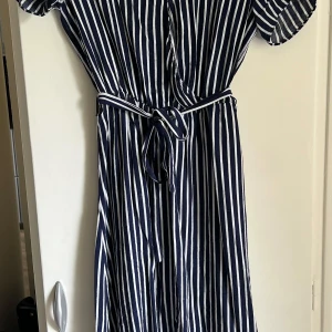 Marinblå och vit randig klänning från Vila - Säljer en marinblå och vit randig klänning från Vila med knytskärp i midjan. Klänningen har korta ärmar och v-ringning, perfekt för en fräsch och stilren look. Lätt och luftig modell som passar till många tillfällen. Använd men i fint skick. Lite skavanker (se bilder) men inget som syns när den är på eller stör funktionen 