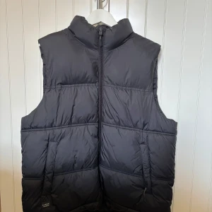 Svart dunväst från Zara - Säljer en svart dunväst från Zara med hög krage och dragkedja framtill. Västen har två fickor med dragkedja och en stilren, quiltad design. Perfekt för lager-på-lager under kyliga dagar. Sparsamt använd.