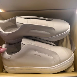 Vita sneakers från Axel Arigato - Säljer ett par stilrena vita sneakers från Axel Arigato. Skorna har minimalistisk design utan snörning, med diskret logga på sidan och rund tå. Perfekta för dig som gillar clean och modern stil. Skorna är aldrig använda säljs på grund av storlek, ordinarie pris ligger runt 2500