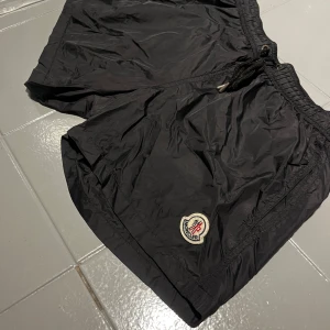 Svarta shorts från Moncler med dragkedja - Svarta shorts från Moncler med elastisk midja och snörning. Shortsen har en dragkedjeficka med röd och vit detalj samt Moncler-logga framtill. Tillverkade i lätt syntetmaterial med meshfoder, perfekta för sommaren. DEM HAR ETT LITET HÅL BREDVID BAK ETIKETTEN PP BILD 4!!!Pris kan diskuteras!