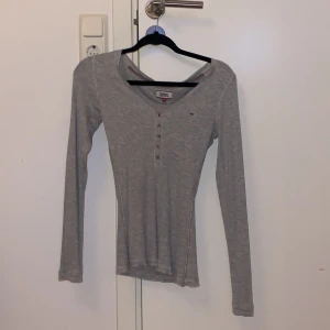 Grå långärmad topp från Tommy Jeans - Säljer en grå långärmad topp från Tommy Jeans med v-ringning och knappar framtill. Tröjan har diskret logga på bröstet och fina röda och blå detaljer längs ärmsömmarna. Ribbad bomullskvalitet som är mjuk och skön.