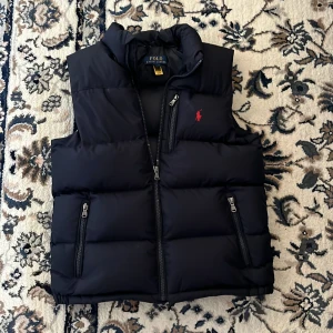 Svart dunväst från Polo Ralph Lauren - Säljer en svart dunväst från Polo Ralph Lauren med hög krage och dragkedja framtill. Västen har tre fickor med dragkedja och en röd broderad logga på bröstet. Perfekt för lager-på-lager-stil.