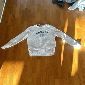 Vit sweatshirt från Morris - Säljer en vit sweatshirt från Morris med blå text och emblem på bröstet. Tröjan har rund halsringning och långa ärmar. Perfekt för en avslappnad och stilren look.
