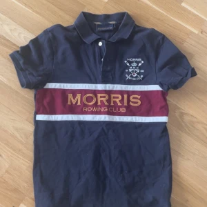 Mörkblå pikétröja från Morris Rowing Club - Säljer en mörkblå pikétröja från Morris Rowing Club med vit och vinröd rand över bröstet och stor gul text. Tröjan har klassisk krage, korta ärmar och broderad logga på bröstet. Perfekt för en sportig och stilren look.