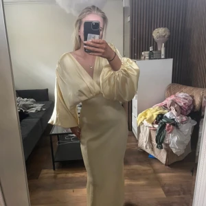 Ljusgul satin långklänning från Flan - Elegant ljusgul långklänning i satin från Flan. Klänningen har djup v-ringning, långa puffärmar och öppen rygg. Materialet är glansigt och följsamt, vilket ger en lyxig känsla. Perfekt för speciella tillfällen. Aldrig använd 