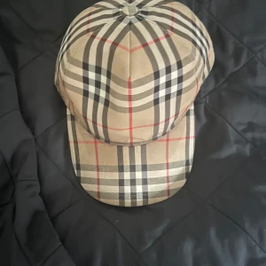  keps från Burberry - Säljer en klassisk rutig keps från Burberry med beige, svarta, vita och röda ränder. Kepsen har en böjd skärm och justerbar baksida. Märket 'BURBERRY LONDON ENGLAND' syns tydligt bak. Perfekt accessoar för en trendig look.