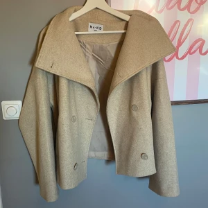 Beige jacka från NA-KD - Säljer en stilren beige jacka från NA-KD med bred krage och dubbelknäppning. Jackan har en kortare passform och är tillverkad i ett ull-liknande material. Använd en gång men som ny. 