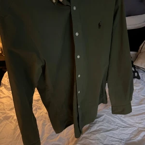 Olivgrön skjorta från Ralph Lauren - Snygg olivgrön skjorta från Ralph Lauren i slim fit-modell. Skjortan är tillverkad i stretchig bomull och har långa ärmar, knappar framtill och klassisk broderad logga på bröstet. Hör av er vid minsta fundering!